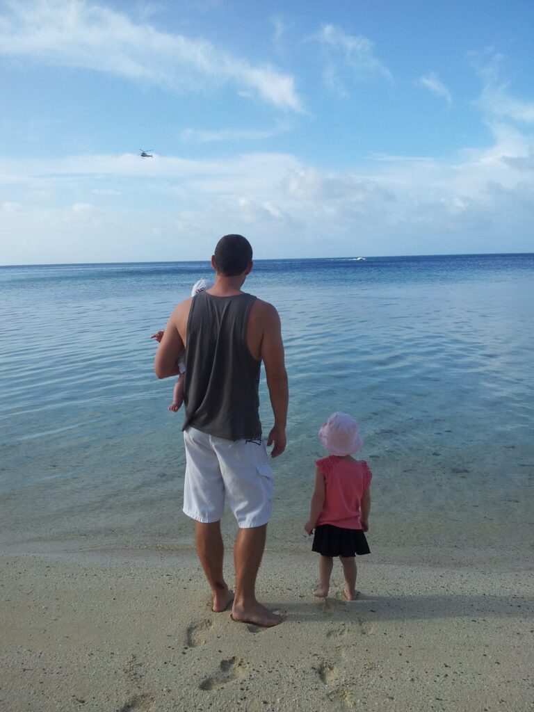 A father and two young children looking out at a calm turquoise ocean, backs to the camera